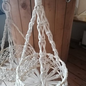 May include: Two white macrame plant hangers with wooden rings. The hangers are made of thick white rope and have a circular base.