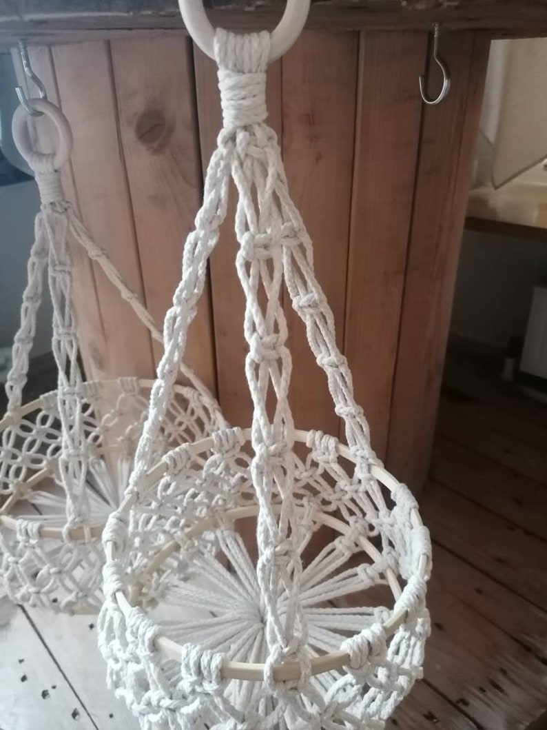 May include: Two white macrame plant hangers with wooden rings. The hangers are made of thick white rope and have a circular base.
