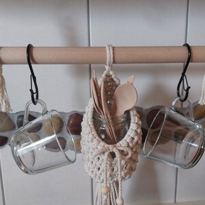 May include: A wooden rod with two black hooks holding two clear glass mugs. A macrame hanging with wooden spoons and a glass jar is hanging from the rod.