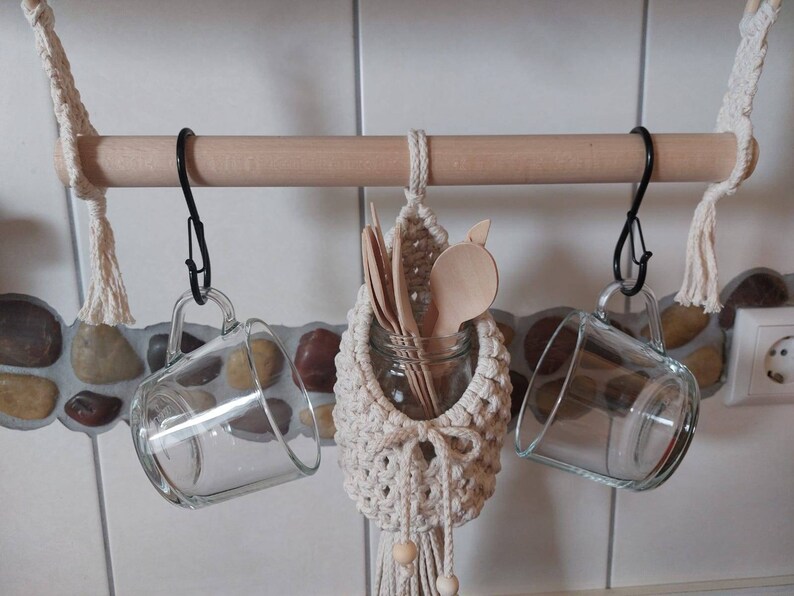 May include: A wooden rod with two black hooks holding two clear glass mugs. A macrame hanging with wooden spoons and a glass jar is hanging from the rod.