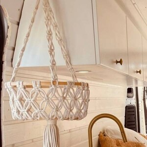Puede incluir: Un macetero colgante de macram&eacute; blanco con un anillo de madera y un borla. El macetero est&aacute; colgado del techo de una furgoneta blanca. Hay un grifo dorado y una almohada marr&oacute;n en el fondo.