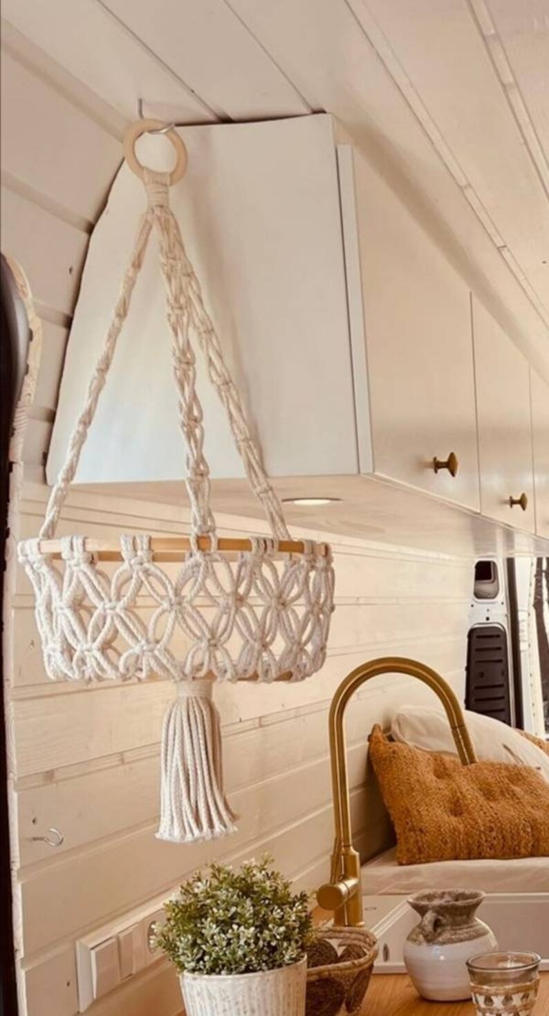 Puede incluir: Un macetero colgante de macram&eacute; blanco con un anillo de madera y un borla. El macetero est&aacute; colgado del techo de una furgoneta blanca. Hay un grifo dorado y una almohada marr&oacute;n en el fondo.