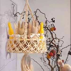 May include: A macrame hanging basket with a wooden ring at the top and a tassel at the bottom. The basket is filled with five stuffed bunny toys in yellow, brown, and white.