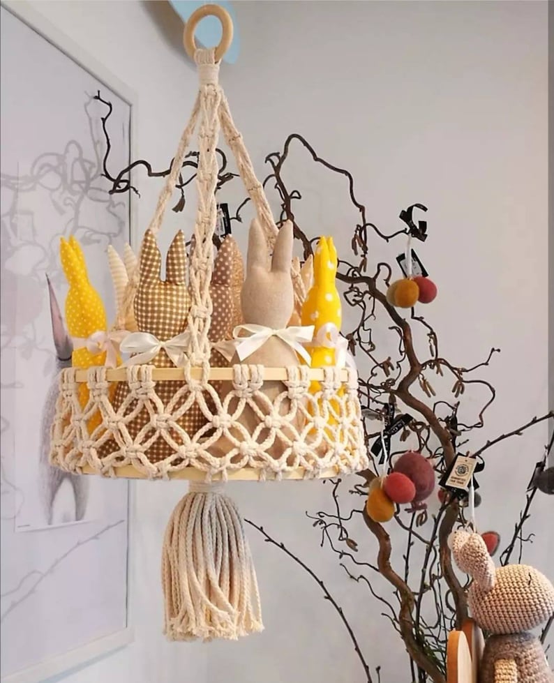 May include: A macrame hanging basket with a wooden ring at the top and a tassel at the bottom. The basket is filled with five stuffed bunny toys in yellow, brown, and white.