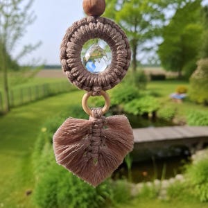 Könnte beinhalten: Ein handgefertigter Makramee-Sonnenfänger mit einem klaren Kristallkugel, die in einem gewebten braunen Ring hängt. Unter dem Ring befindet sich ein blattförmiges Makramee-Design. Der Sonnenfänger hängt an einer braunen Kordel und einer Holzperle. Verschwommener Hintergrund.