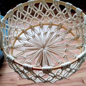 May include: A white macrame hanging basket with a wooden frame. The basket is made with a knotting technique that creates a circular pattern.
