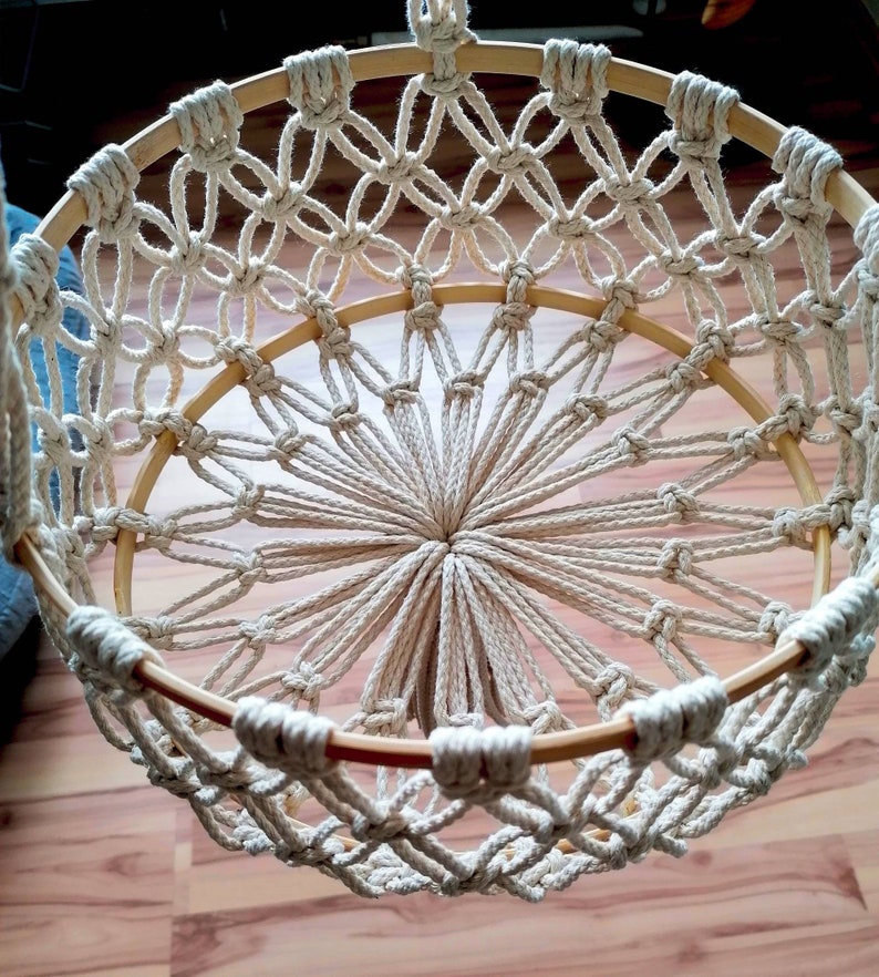 May include: A white macrame hanging basket with a wooden frame. The basket is made with a knotting technique that creates a circular pattern.