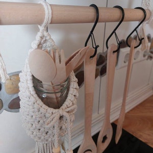May include: A white macrame hanging organizer with a wooden bead and a glass jar for storing utensils. The organizer is hanging from a wooden rod with black metal hooks. There are wooden spoons and spatulas hanging from the hooks.