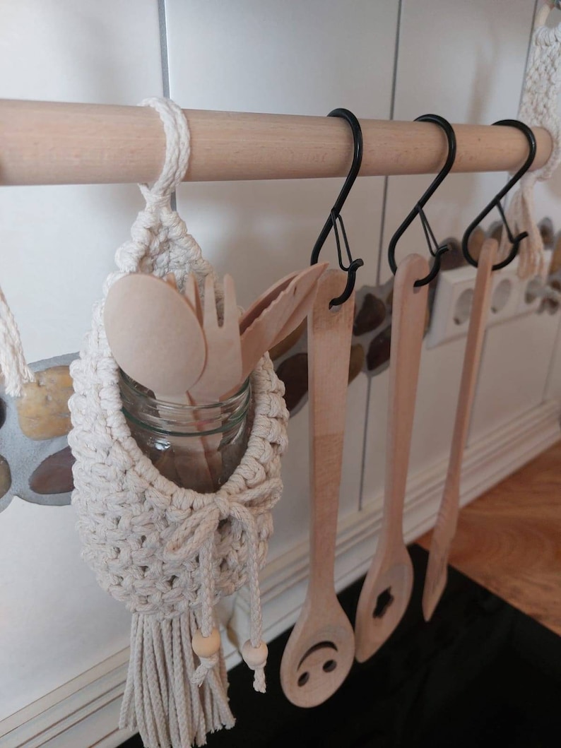 May include: A white macrame hanging organizer with a wooden bead and a glass jar for storing utensils. The organizer is hanging from a wooden rod with black metal hooks. There are wooden spoons and spatulas hanging from the hooks.