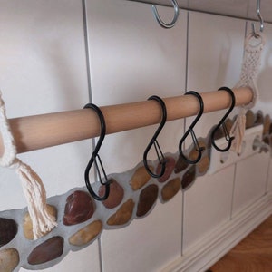 May include: A wooden rod with four black metal hooks attached to it. The rod is mounted to a white tiled wall with a decorative stone border.