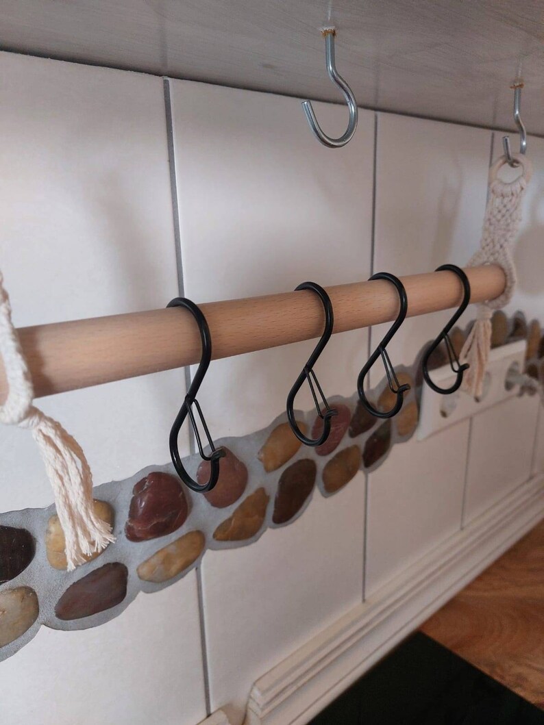 May include: A wooden rod with four black metal hooks attached to it. The rod is mounted to a white tiled wall with a decorative stone border.