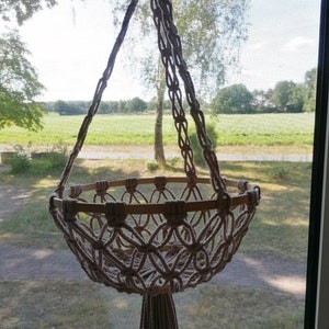 Puede incluir: Un macetero de macram&eacute; con un anillo de madera y un borla. El macetero est&aacute; hecho de cuerda de algod&oacute;n beige natural y est&aacute; colgado frente a una ventana.