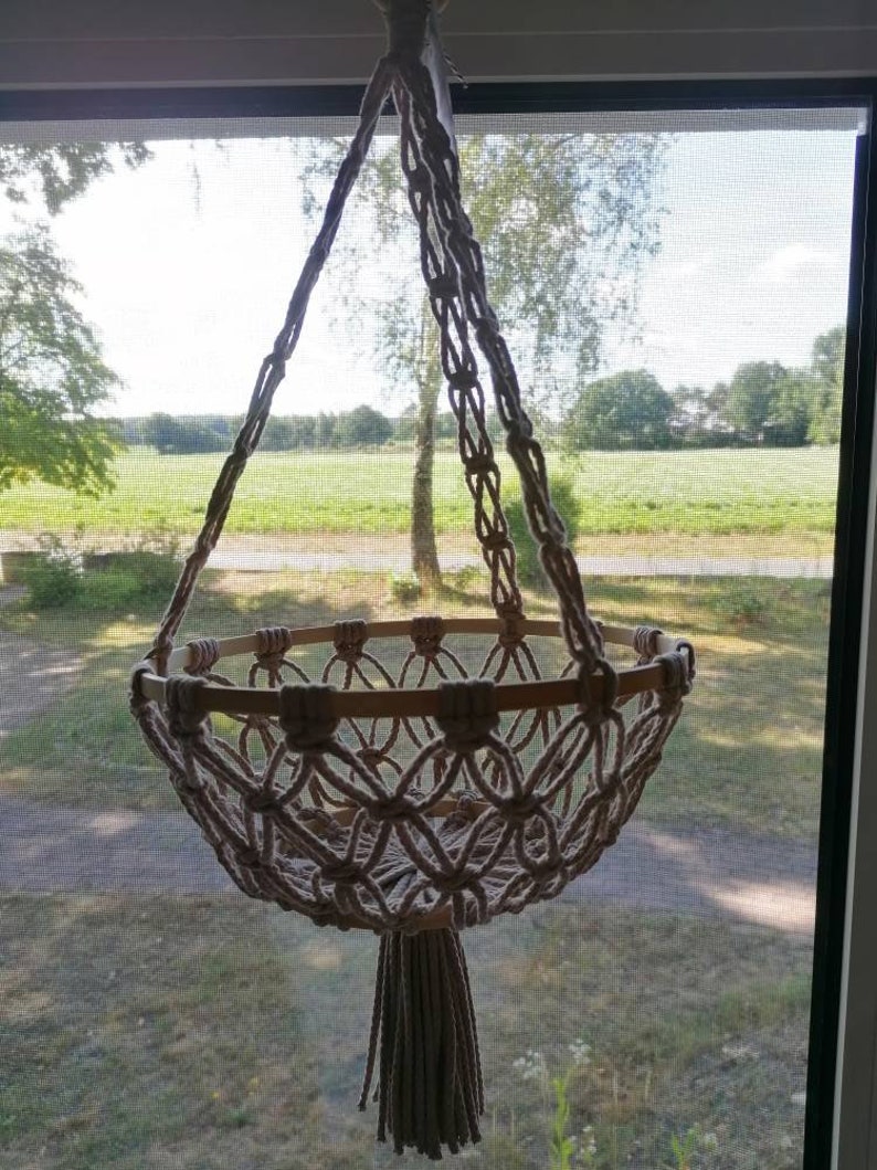 Puede incluir: Un macetero de macram&eacute; con un anillo de madera y un borla. El macetero est&aacute; hecho de cuerda de algod&oacute;n beige natural y est&aacute; colgado frente a una ventana.