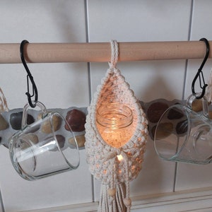 May include: A wooden rod with three hanging items: a glass mug, a glass mug with a crocheted macrame holder, and a glass jar with a candle inside the crocheted macrame holder.
