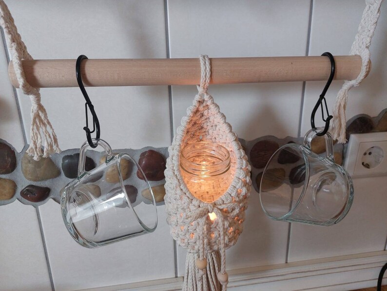 May include: A wooden rod with three hanging items: a glass mug, a glass mug with a crocheted macrame holder, and a glass jar with a candle inside the crocheted macrame holder.
