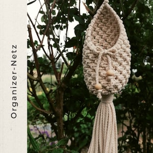 May include: A cream-coloured macrame hanging organiser with a woven basket design, a tassel, and wooden beads. The text "Organizer-Netz" is printed vertically on the left side of the image. The organiser is suspended from a tree branch.