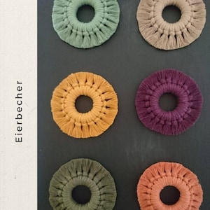 Op de afbeelding: Zes handgemaakte macramé-onderzetters in verschillende kleuren, waaronder groen, beige, geel, paars en oranje. Elke onderzetter heeft een cirkelvormig ontwerp met een centraal gat en een franje rand.