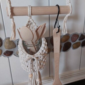 May include: A wooden dowel rod with a hanging macrame utensil holder with a glass jar and wooden utensils inside. A wooden spoon with a smiley face is hanging from a hook on the rod.