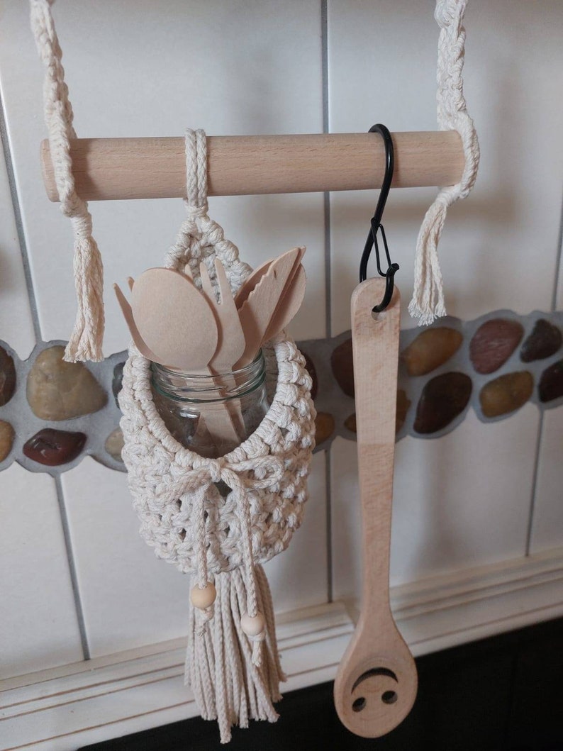 May include: A wooden dowel rod with a hanging macrame utensil holder with a glass jar and wooden utensils inside. A wooden spoon with a smiley face is hanging from a hook on the rod.