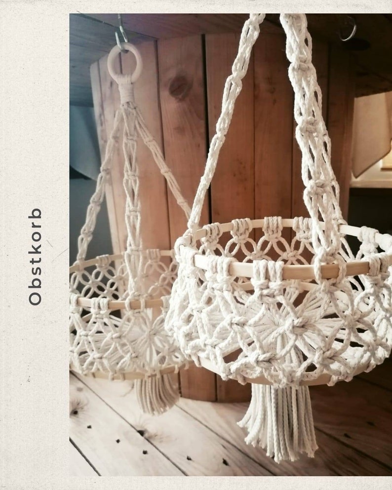 May include: Two macrame hanging baskets with wooden rings and tassels. The baskets are made of natural white cotton rope and are hanging from a wooden surface. The text "Obstkorb" is visible in the image.