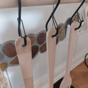 May include: Three wooden kitchen utensils hanging on black S-shaped hooks attached to a wooden bar. The utensils are a wooden spoon, a wooden spatula, and a wooden ladle.
