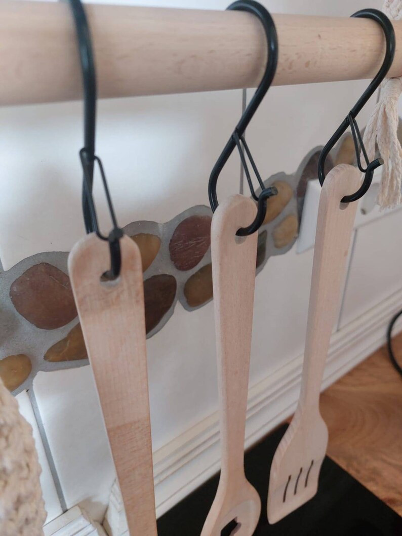 May include: Three wooden kitchen utensils hanging on black S-shaped hooks attached to a wooden bar. The utensils are a wooden spoon, a wooden spatula, and a wooden ladle.