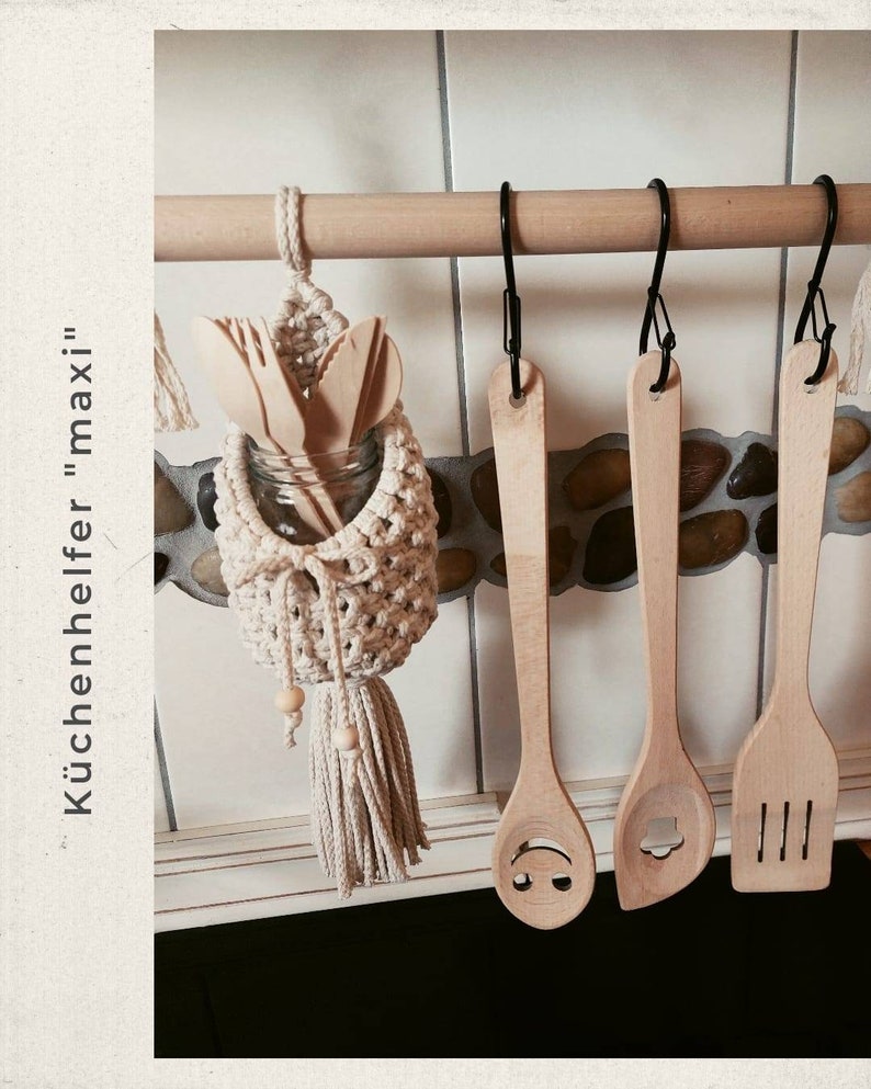 May include: A white crocheted hanging utensil holder with a wooden dowel and wooden spoons hanging from it. The holder has a tassel and beads. The text "K&uuml;chenhelfer "maxi" " is visible in the image.