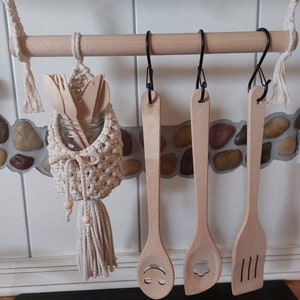 May include: Wooden kitchen utensils hanging on a wooden rack with a macrame utensil holder. The utensils include a wooden spoon with a smiley face carved into it, a wooden spoon with a flower carved into it, and a wooden spatula with three lines carved into it.