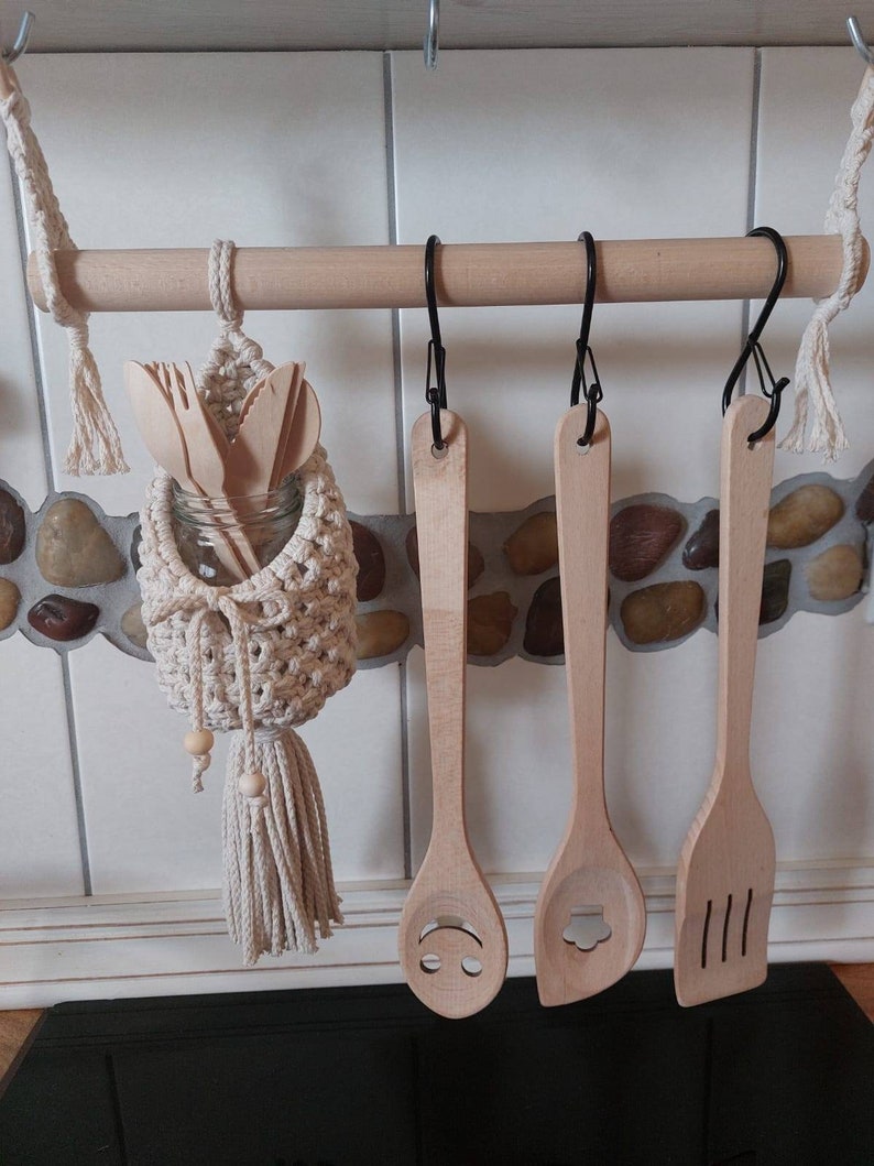 May include: Wooden kitchen utensils hanging on a wooden rack with a macrame utensil holder. The utensils include a wooden spoon with a smiley face carved into it, a wooden spoon with a flower carved into it, and a wooden spatula with three lines carved into it.