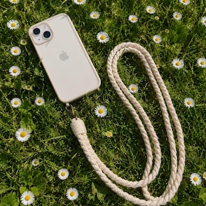 Könnte beinhalten: Ein helles Smartphone in einer Schutzhülle, das an einem langen, dicken, cremefarbenen geflochtenen Seilband befestigt ist. Das Band verfügt über einen bronzefarbenen Metallverschluss, der das Seil mit der Unterseite der Handyhülle verbindet und das Tragen des Geräts ermöglicht.