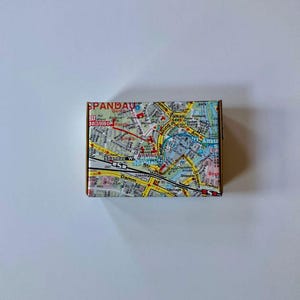 Peut inclure: Une petite boîte en carton avec une carte de Berlin, Allemagne, imprimée sur le dessus. La carte montre les principales rues et les monuments de la ville, notamment la Porte de Brandebourg, le Reichstag et le Mémorial du mur de Berlin.