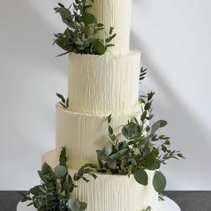 Puede incluir: Una tarta de boda de cuatro pisos con glaseado blanco texturizado y acentos de follaje verde fresco. La tarta se asienta sobre un soporte blanco, con cada capa disminuyendo de tamaño. Decorada con hojas verdes.