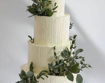 May include: A four-tiered wedding cake with textured white frosting and fresh green foliage accents. The cake sits on a white cake stand, with each layer decreasing in size as it rises. The cake is decorated with green leaves.