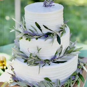 Könnte beinhalten: Eine dreistöckige weiße Hochzeitstorte, die mit Lavendelzweigen und Olivenzweigen verziert ist. Die Torte steht auf einem Holzkuchenständer mit den Initialen "B & M", die in das Holz geschnitzt sind.