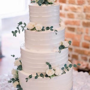 Peut inclure: Un gâteau de mariage blanc à quatre étages décoré de roses blanches et de feuillage d'eucalyptus.