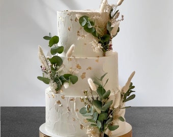 May include: A three-tiered white wedding cake decorated with gold leaf and greenery. The cake is adorned with sprigs of eucalyptus, dried pampas grass, and small white flowers.