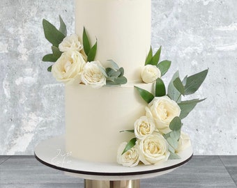 May include: A two-tiered white cake adorned with white roses and green foliage. The cake is presented on a white cake stand with a gold-coloured base. The cake is decorated with fresh flowers and greenery.