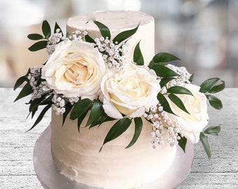 Könnte beinhalten: Zweistöckige weiße Hochzeitstorte, verziert mit weißen Rosen und Grün.