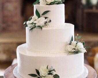 May include: A three-tiered white wedding cake decorated with white flowers and green leaves.