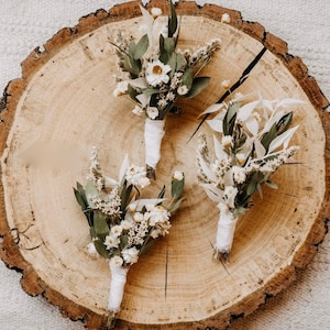 Puede incluir: Tres pequeños ramos de flores blancas secas y vegetación dispuestos sobre una rodaja de madera. Los ramos están atados con una cinta blanca.