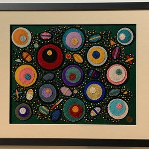 May include: A framed artwork featuring a green background with colorful circles and other shapes sewn onto the fabric. The circles are various sizes and colors, including red, yellow, blue, pink, and white.