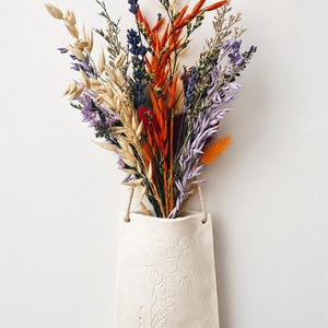 May include: A wall-mounted white ceramic pocket vase featuring an embossed floral design on the front. The vase holds a dried flower arrangement containing purple lavender, orange wheat stalks, light purple grasses, and various green foliage.