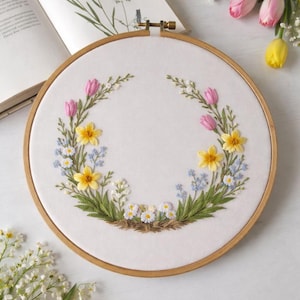 May include: A circular embroidery hoop with a floral design. The design includes pink tulips, yellow daffodils, and small white and blue flowers arranged in a wreath. The hoop is wooden and the fabric is white.