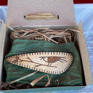May include: A wooden hair clip with a bird design, presented in a cardboard gift box with green tissue paper and shredded paper filler. The bird is brown and black, with a branch design.