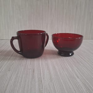 May include: Two ruby red glass cups, one with two handles and the other a bowl with a rounded base.