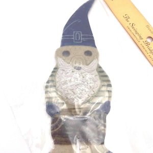 May include: A gnome-shaped ornament in a clear plastic package. The gnome has a blue hat, striped shirt, and a white beard. The gnome's boots and hat trim are blue. A yellow ruler with text is in the background.