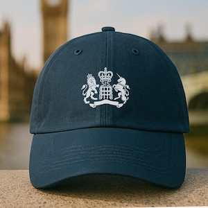 Puede incluir: Gorra de béisbol azul marino con un emblema blanco que presenta un león, un unicornio, una corona y las letras "SIS". La gorra tiene una visera curva y un cierre de correa. El fondo muestra un edificio borroso y agua.