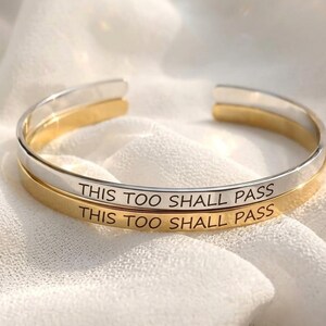 Puede incluir: Dos brazaletes abiertos, uno de tono plateado y otro de tono dorado, cada uno grabado con el texto negro THIS TOO SHALL PASS. Los brazaletes están apilados, con un acabado liso y pulido y un diseño circular simple con una abertura.