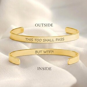 Puede incluir: Dos brazaletes de metal en tono dorado. El brazalete superior presenta el texto grabado "THIS TOO SHALL PASS" en el exterior. El brazalete inferior presenta el texto grabado "BUT WTF?!" en el interior. Ambos brazaletes tienen un acabado liso.