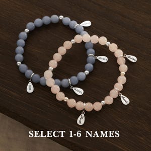 Pode incluir: Duas pulseiras de contas com pingentes de nome personalizados. Uma pulseira tem contas azul-acinzentadas, a outra rosa. Cada pulseira tem detalhes prateados e etiquetas com nomes. O texto na parte inferior diz "SELECT 1-6 NAMES".